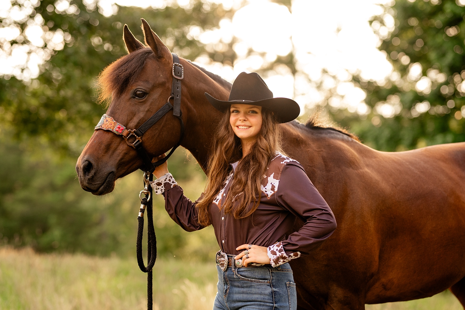 Barrel racer poses with her horse wearing cowgirl hat and cow hide shirt.