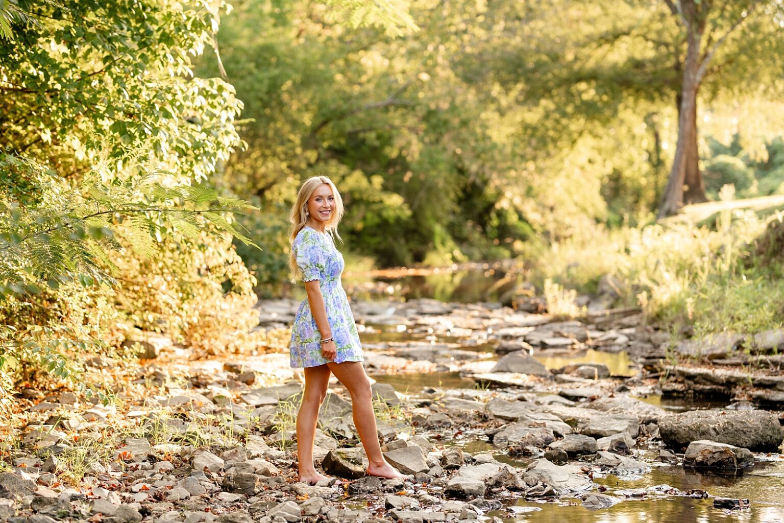 Girl takes senior pictures in pretty purple floral dress. Outfit ideas for senior pictures. Senior photos in Birmingham, senior portraits, creek photoshoot, sunset photography, high school senior photographer