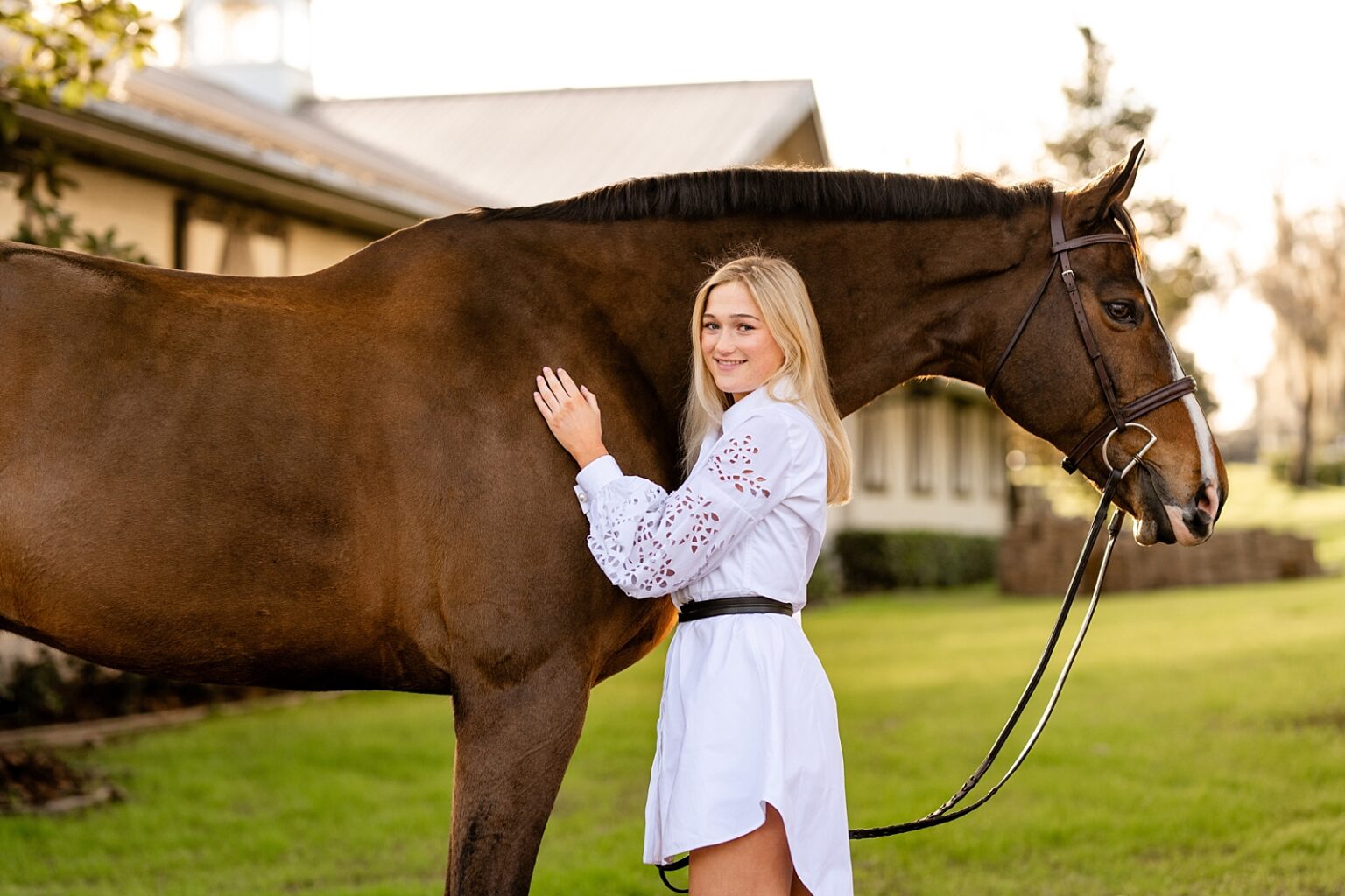 Equestrian inspired senior outfit for senior pictures. White dress with equestrian belt and tall boots. Ocala, FL. Hunter/jumper rider. Golden Oaks Farm LLC. Posing with horses. Sunset.
