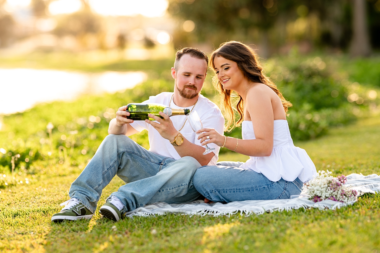 Gracee and Chace sit on a blanket in the grass at golden hour, holding champagne glasses during their Florida engagement session.